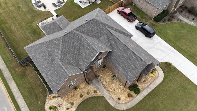 Aerial view of a suburban house with a gray roof, landscaped front yard, and parked cars in the driveway.