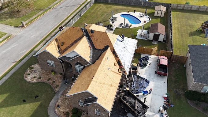 Aerial view of a house with workers installing a new roof. Materials and tools are on the driveway, and there is a pool in the backyard.