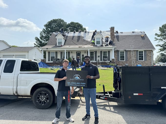 Two people stand in front of a house with roof work in progress. They hold a sign with a business logo and phone number. A truck and trailer are parked nearby.
